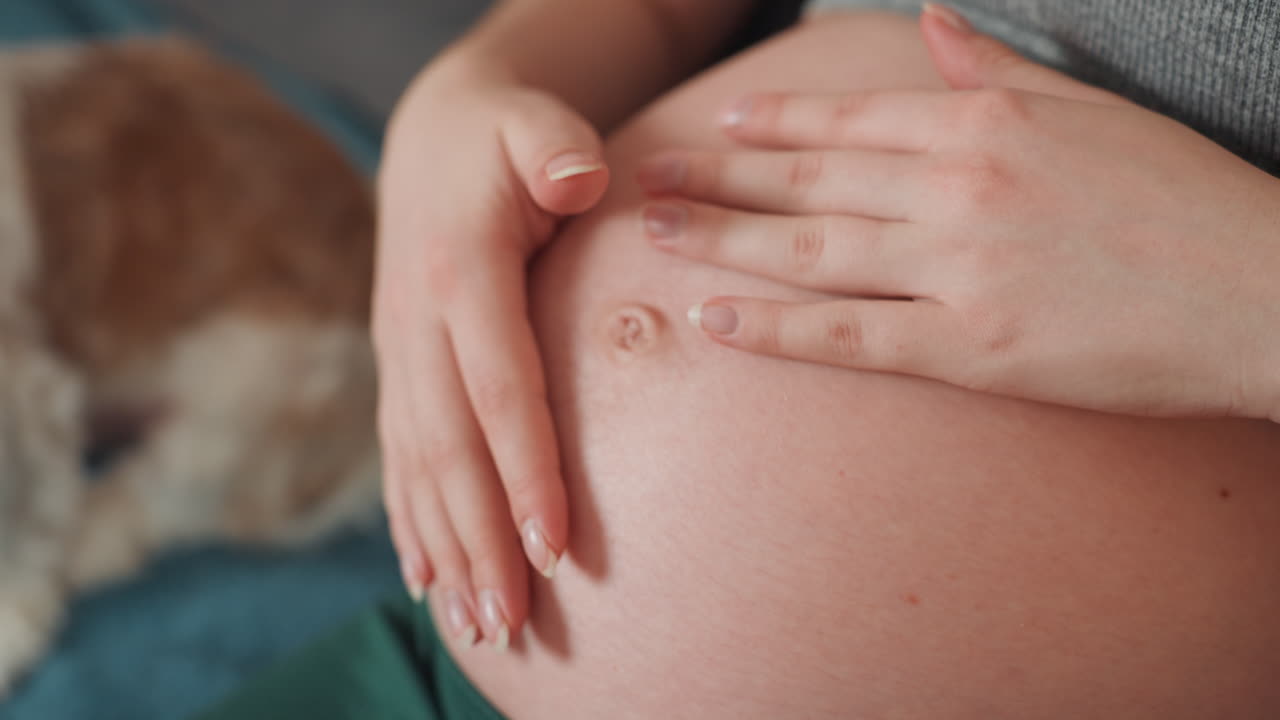 Primo piano della pancia di una donna incinta a casa, madre in attesa che accarezza dolcemente la pancia con unghie smaltate, cane sfocato sullo sfondo, coperta morbida e luce calda, atmosfera tenera e accogliente che mette in risalto la pelle.