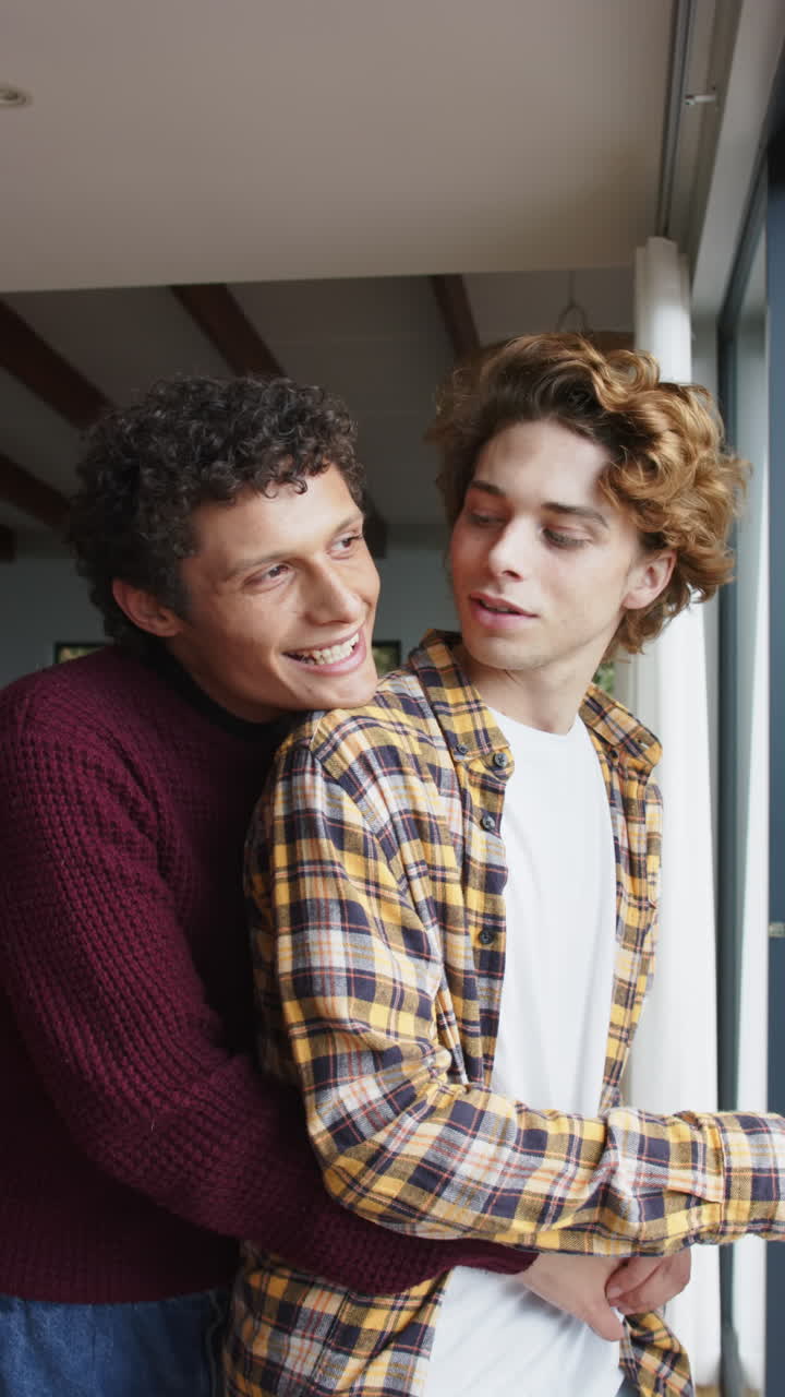 Video vertical de feliz pareja gay diversa de hombres mirando por la ventana en casa, a cámara lenta.