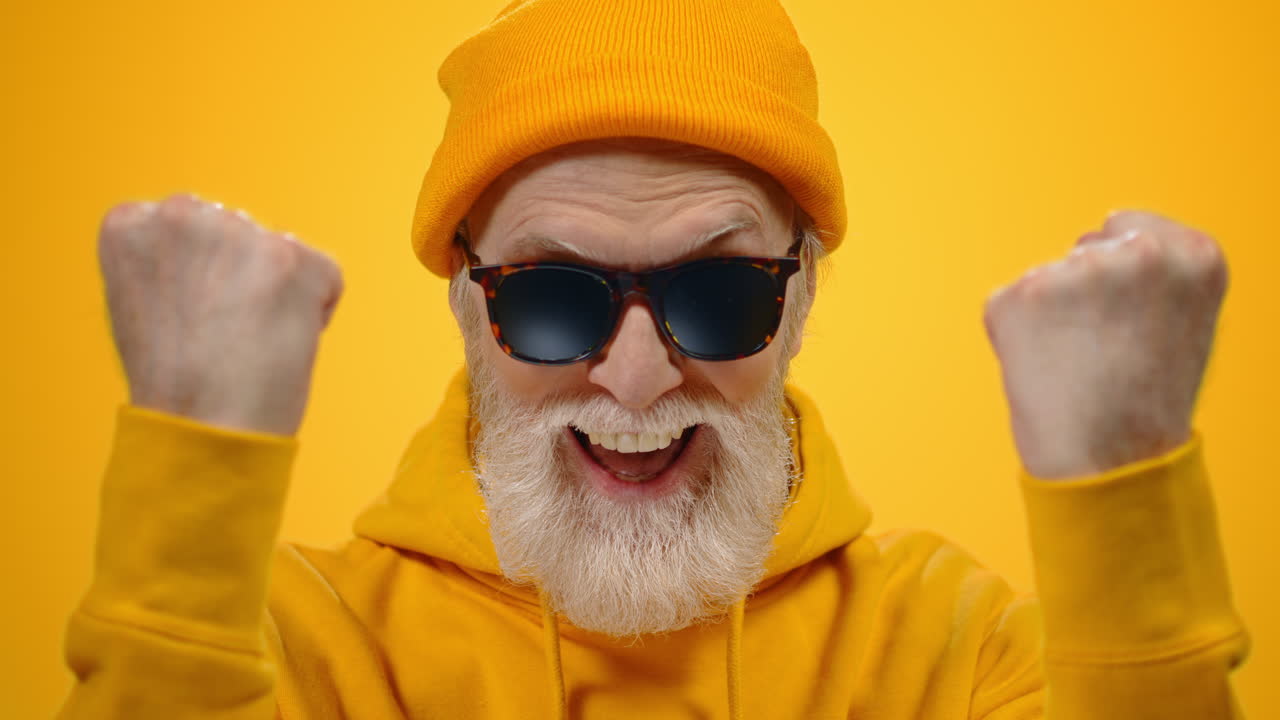 Joyful elderly man smiling in studio. Hipster old guy raising fists indoors.
