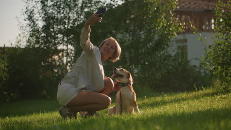 woman taking selfie with her joyful dog outdoors in lush green garden under sunny weather, with building and trees in background, dog is sitting, panting happily as owner crouches beside it