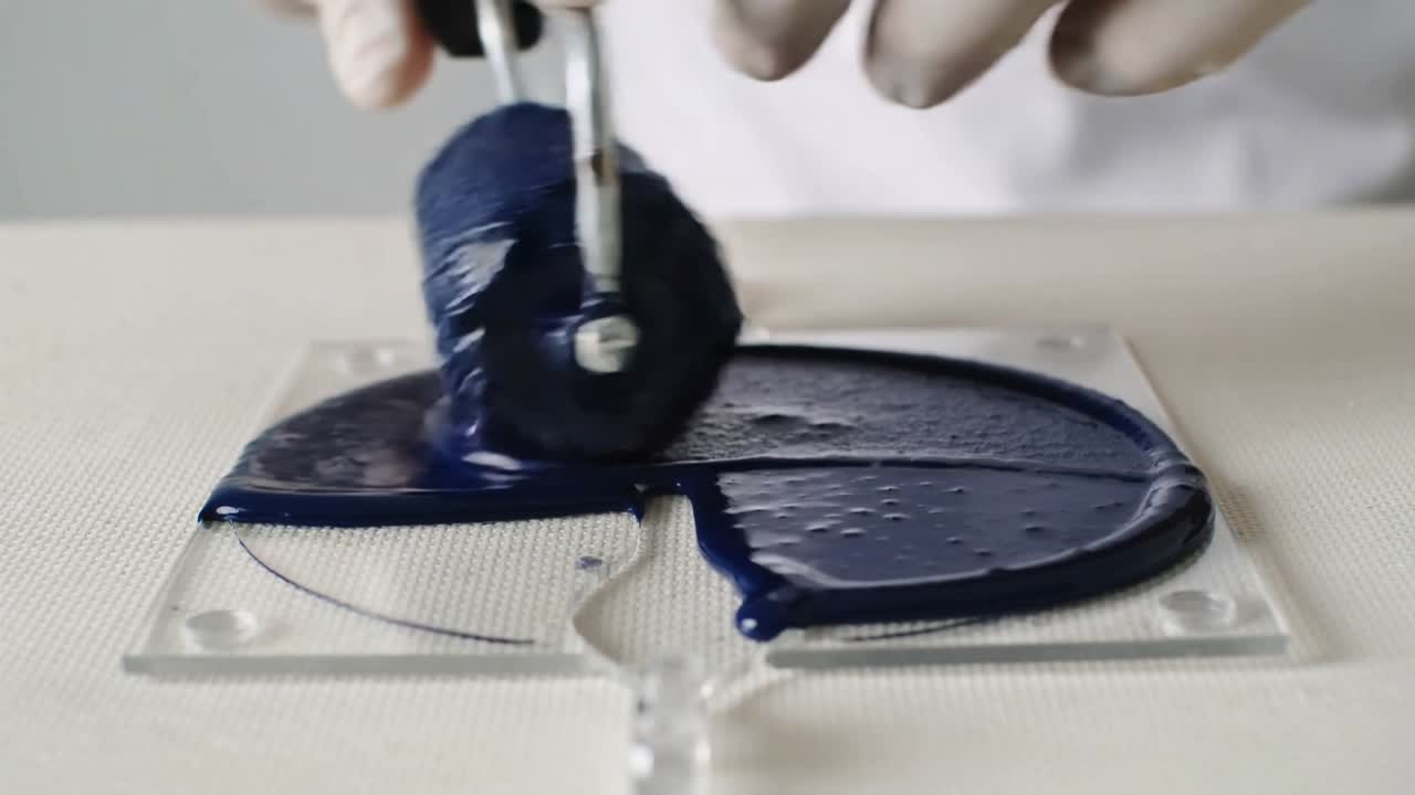 Gloved hands placing loaded brayer rolling across plate on bench, filling round guide with blue ink