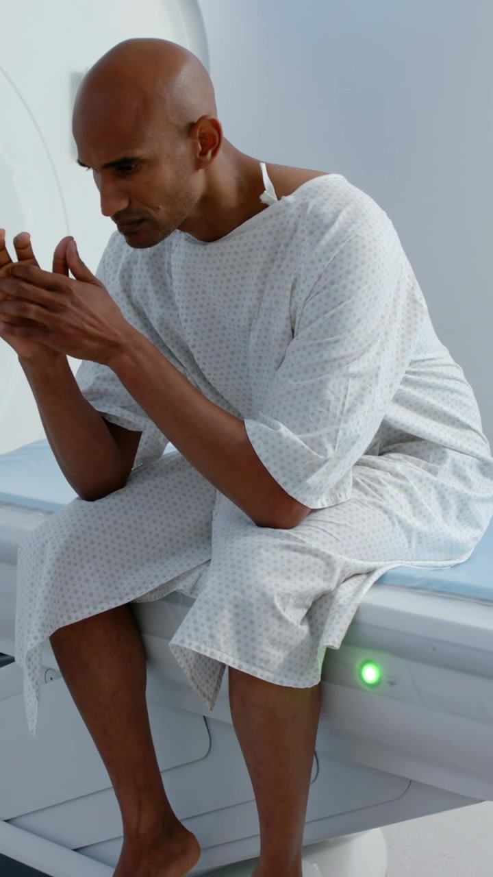 Vertical video: Sitting man in gown fidgeting hands and lifting gaze at scan bed preparing for scan