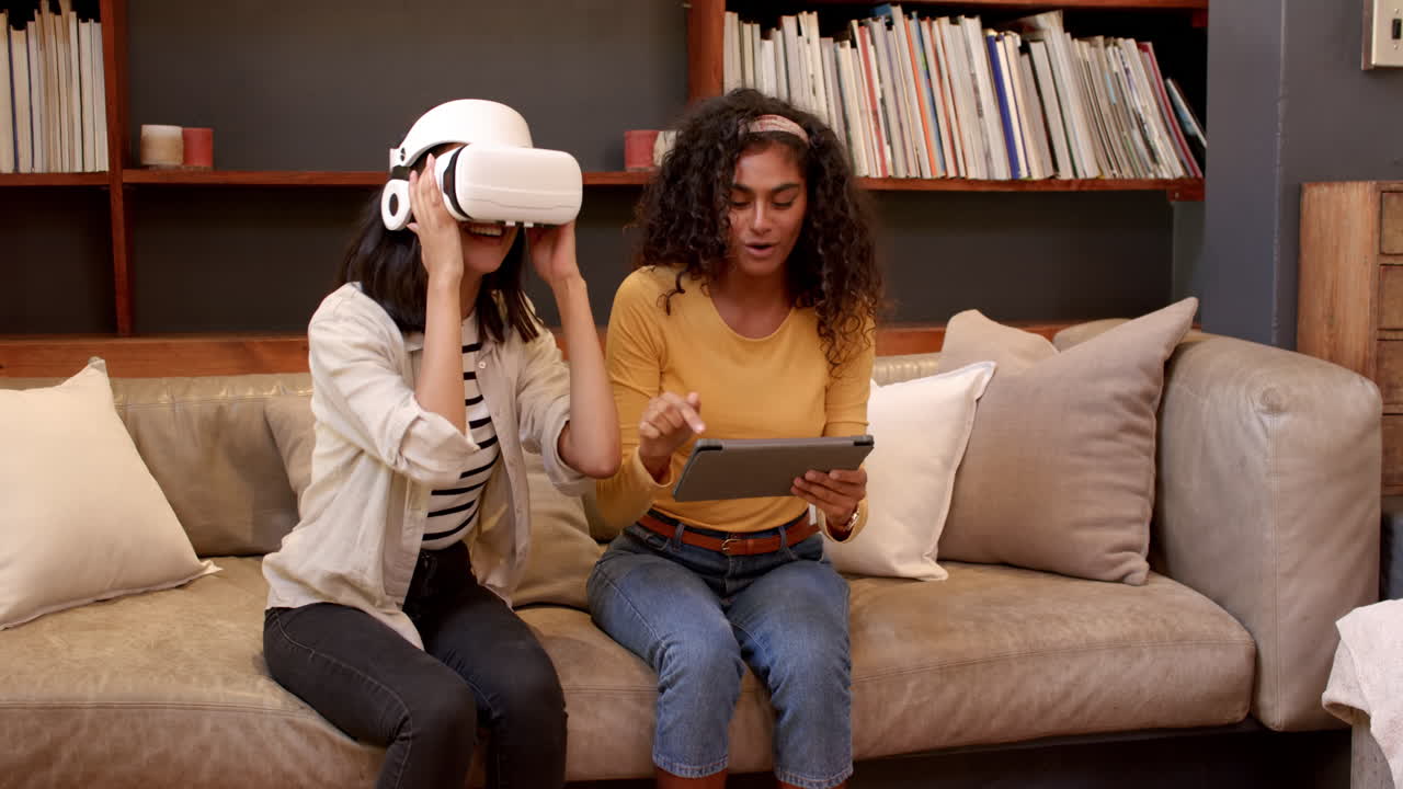 Multiracial female friends enjoying virtual reality and tablet on cozy living room sofa, at home