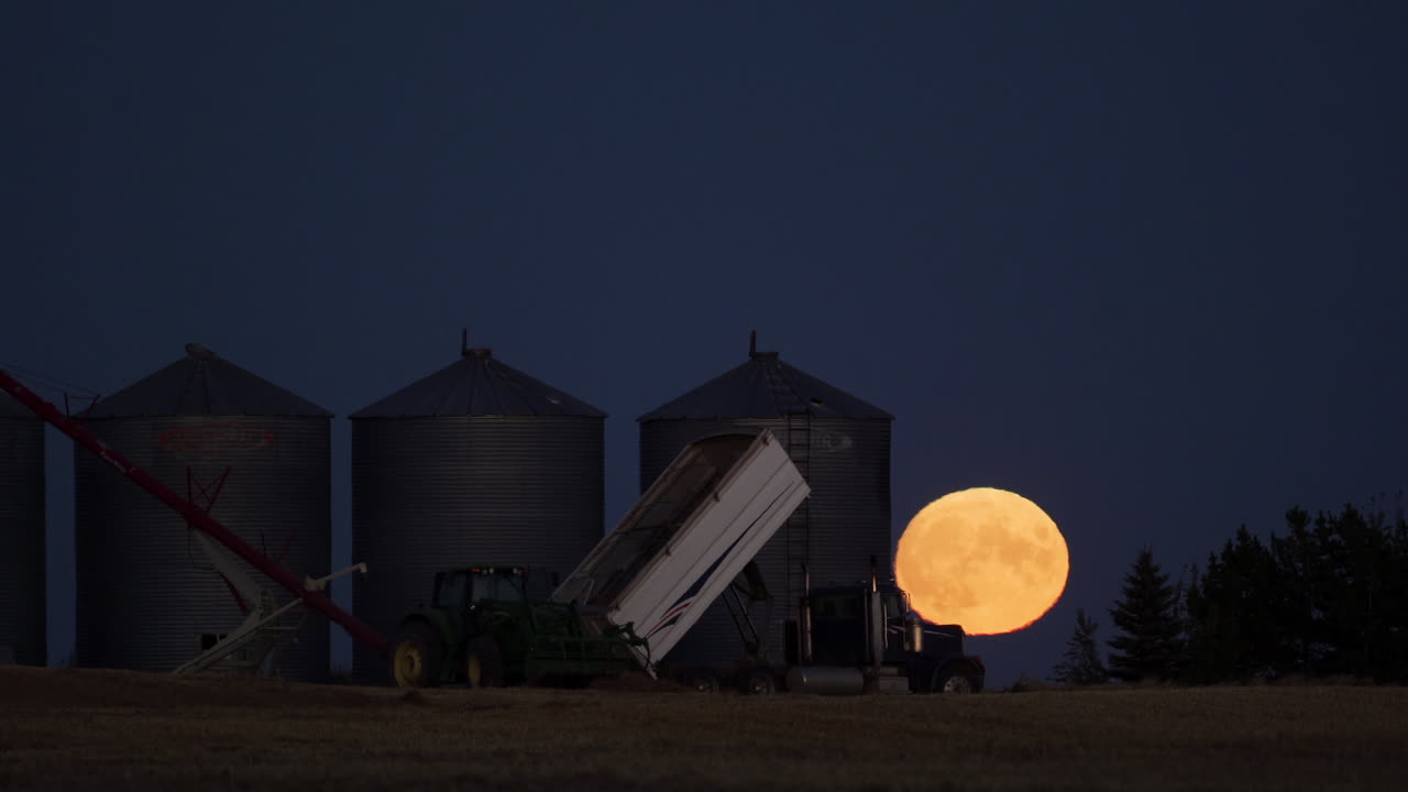 Timelapse teleobjetivo de lua cheia nascendo atrás de silos de grãos durante a colheita