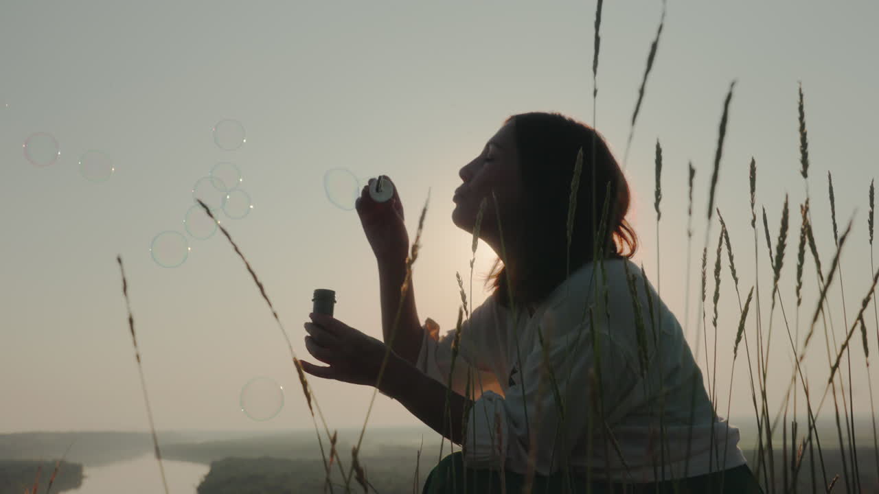 Young Woman blowing bubbles surrounded by tall grain stalks during sunset, silhouette framed by soft golden light and gentle sky, peaceful natural moment as bubbles float upward