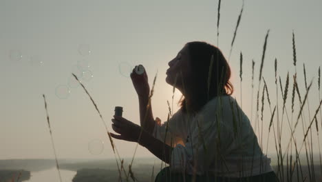 young woman blowing bubbles surrounded by tall grain stalks during sunset, silhouette framed by soft golden light and gentle sky, peaceful natural moment as bubbles float upward