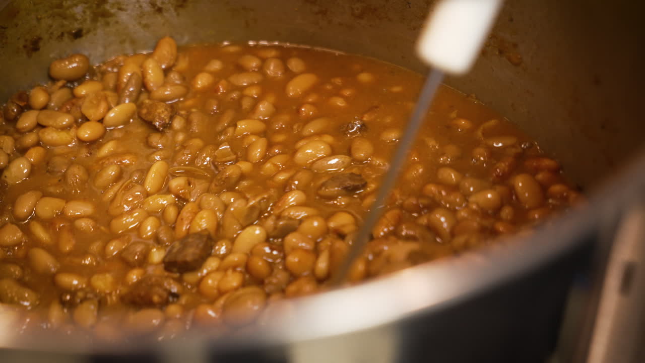 Large spoon stirs huge pot filled with barbecue pinto beans, slow motion close up 4K