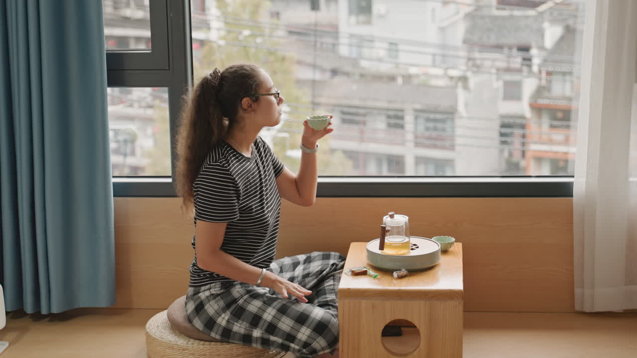 Woman relaxing with snack and beverage. Lady seated near window indulging in drink and light snack. Individual peacefully enjoying beverage and small snack while overlooking urban scenery