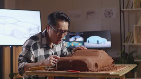 asian man automotive designer works on the sculpture of car clay using rake or wire to smooth out the surface and create details in the design in the studio