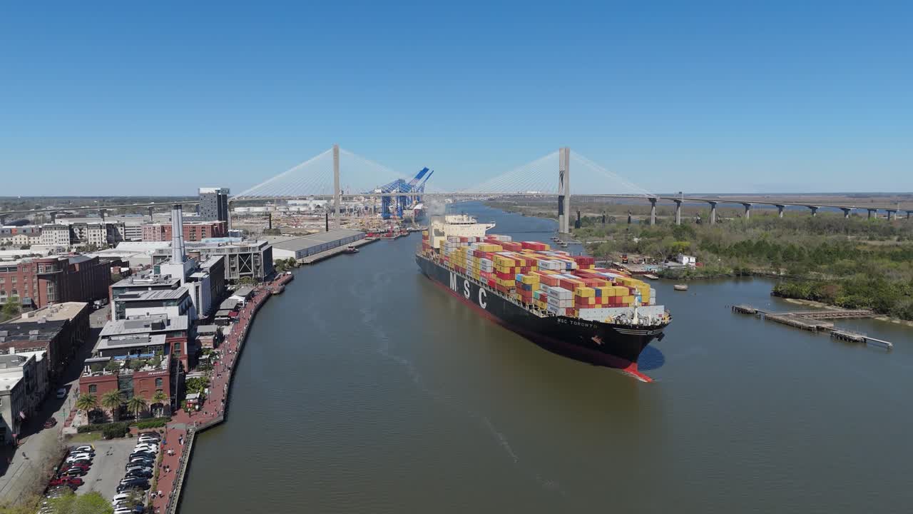Aerial push in on a cargo ship cruising past Savannah's North Historic District with The Talmadge memorial Bridge in the background.