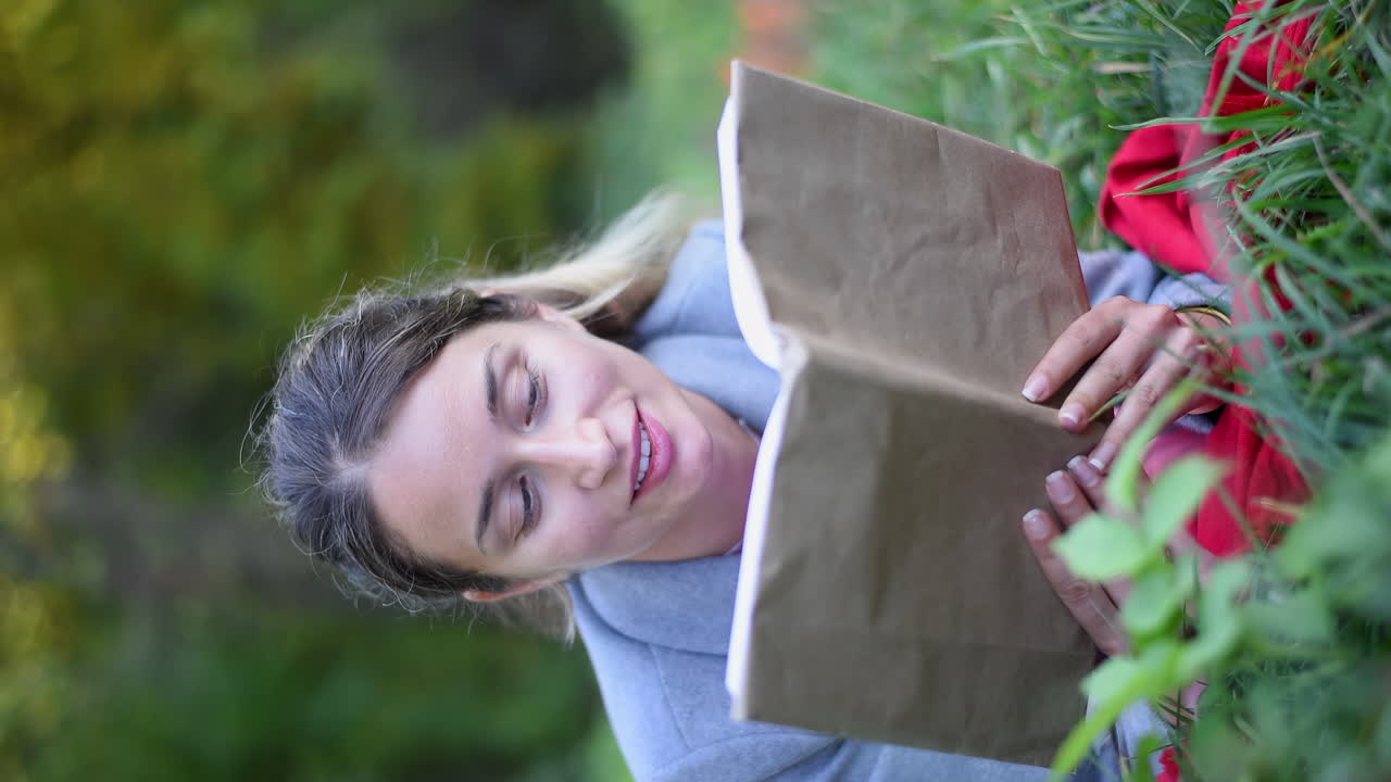 Blonde woman reading a book, lying on the grass in a park. Vertical