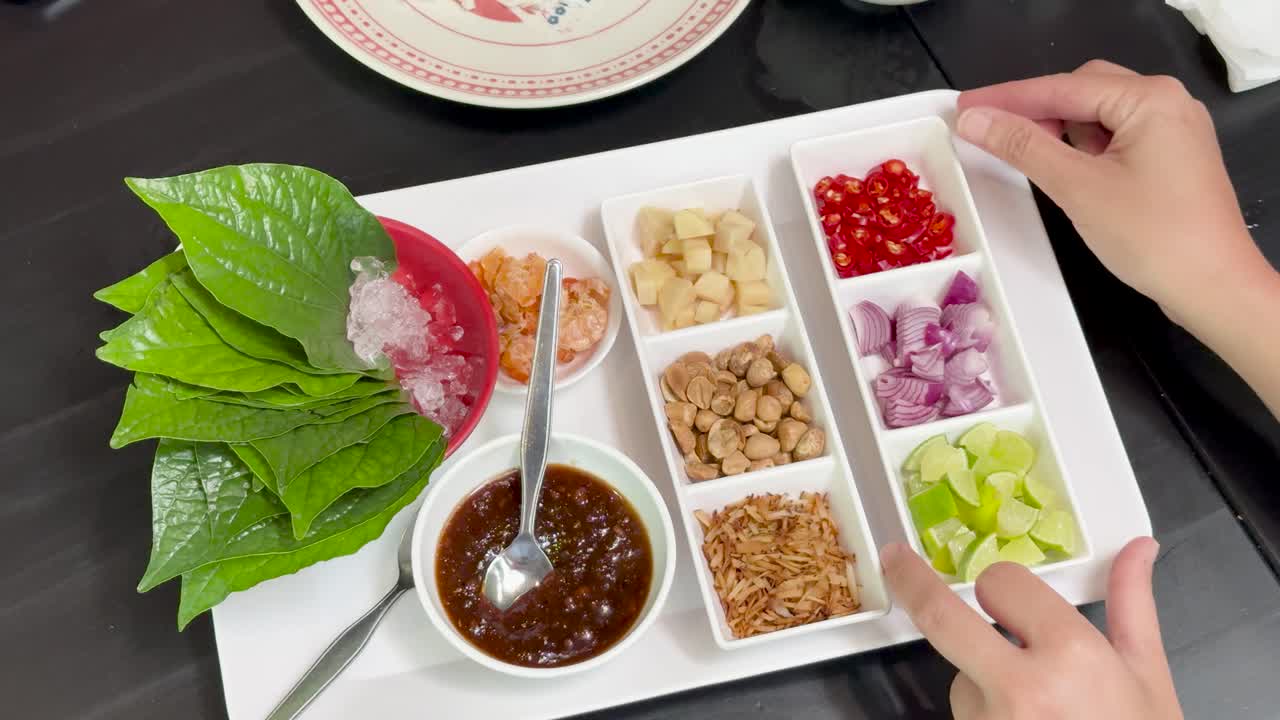 Hands organize colorful Miang Kham ingredients—herbs, peanuts, lime, onion, chili, dried shrimp, and sauce—on a white tray under bright, even lighting, viewed from above