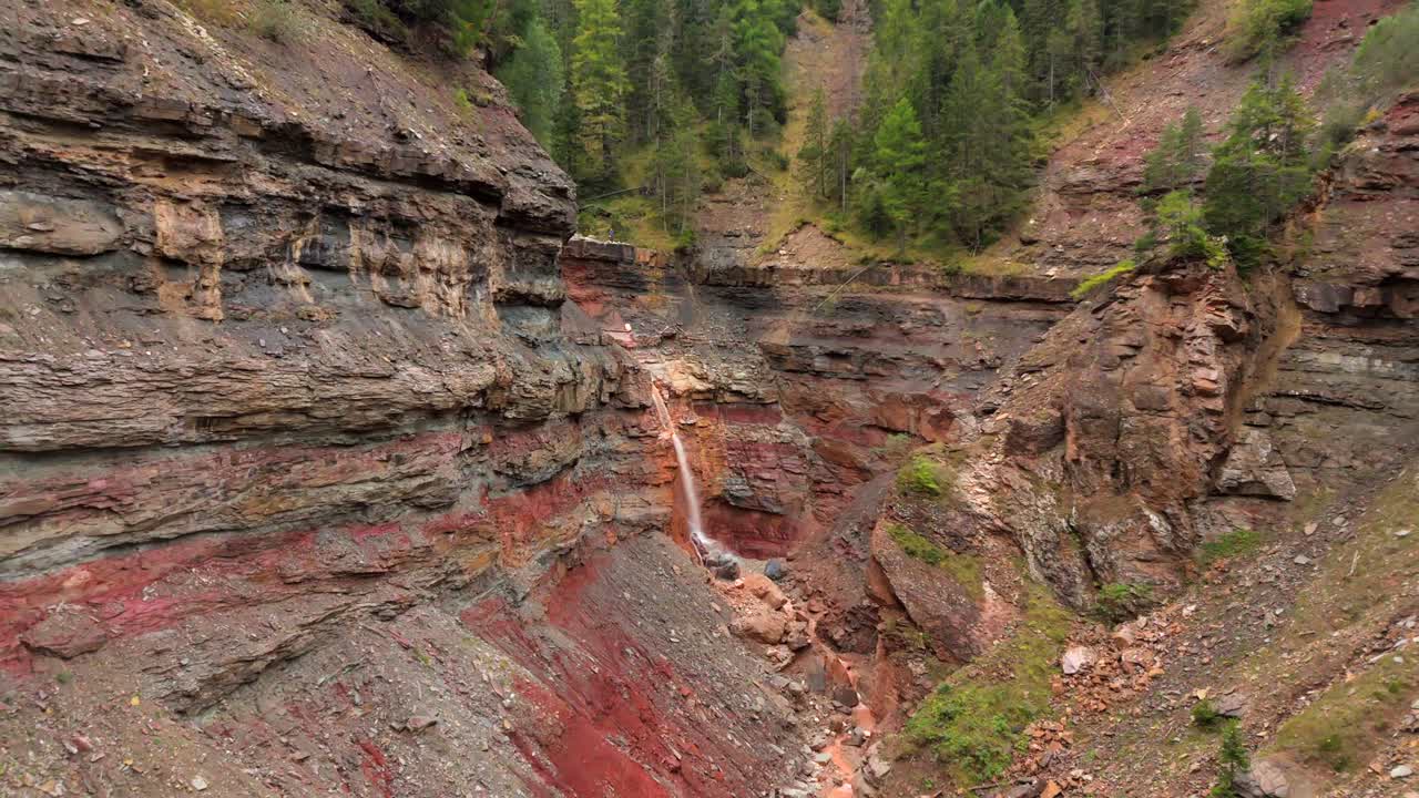 Slow motion clip of a waterfall cascading down colorful, layered rock formations in Bletterbach Gorge, South Tyrol, Italy. The rugged canyon landscape showcases geological layers surrounded by forest.