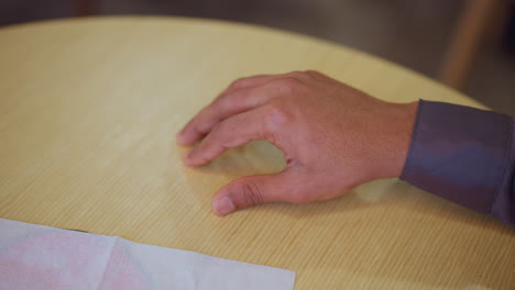 close-up of male hand resting on wooden table surface with relaxed fingers slightly curled, casual positioning suggests pause during conversation or moment of thought in cozy indoor setting with soft lighting
