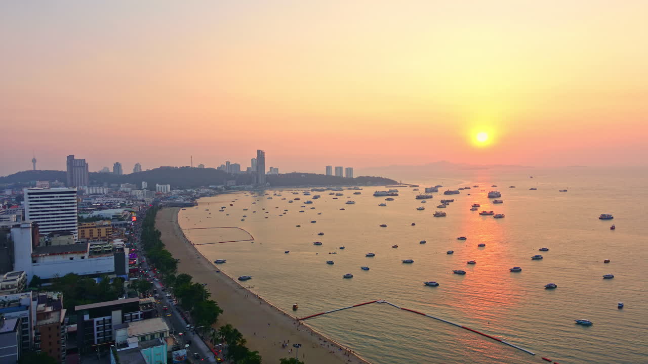 Evening to sunset time lapse view of Pattaya City beach as daylight fades to night. Bright colors in the sky as sun sets on coastal city in Thailand. Beach with boats moving in water and cars on road