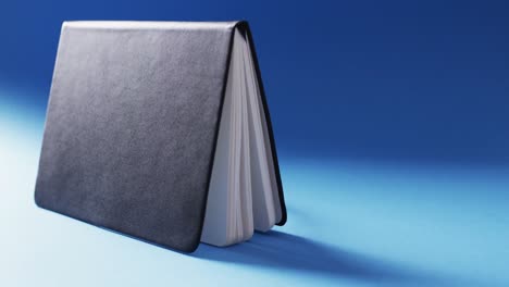 Close-up-of-open-black-book-standing-horizontal-with-copy-space-on-blue-background-in-slow-motion