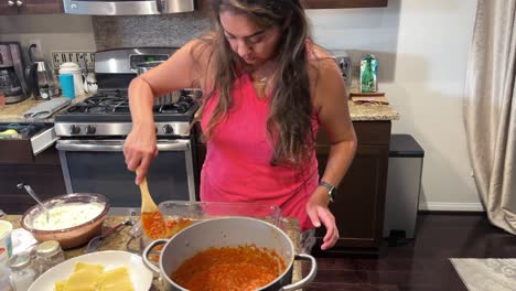 hispanic young woman spreading pasta sauce over lasagna noodles in glass backing dish, in home kitchen