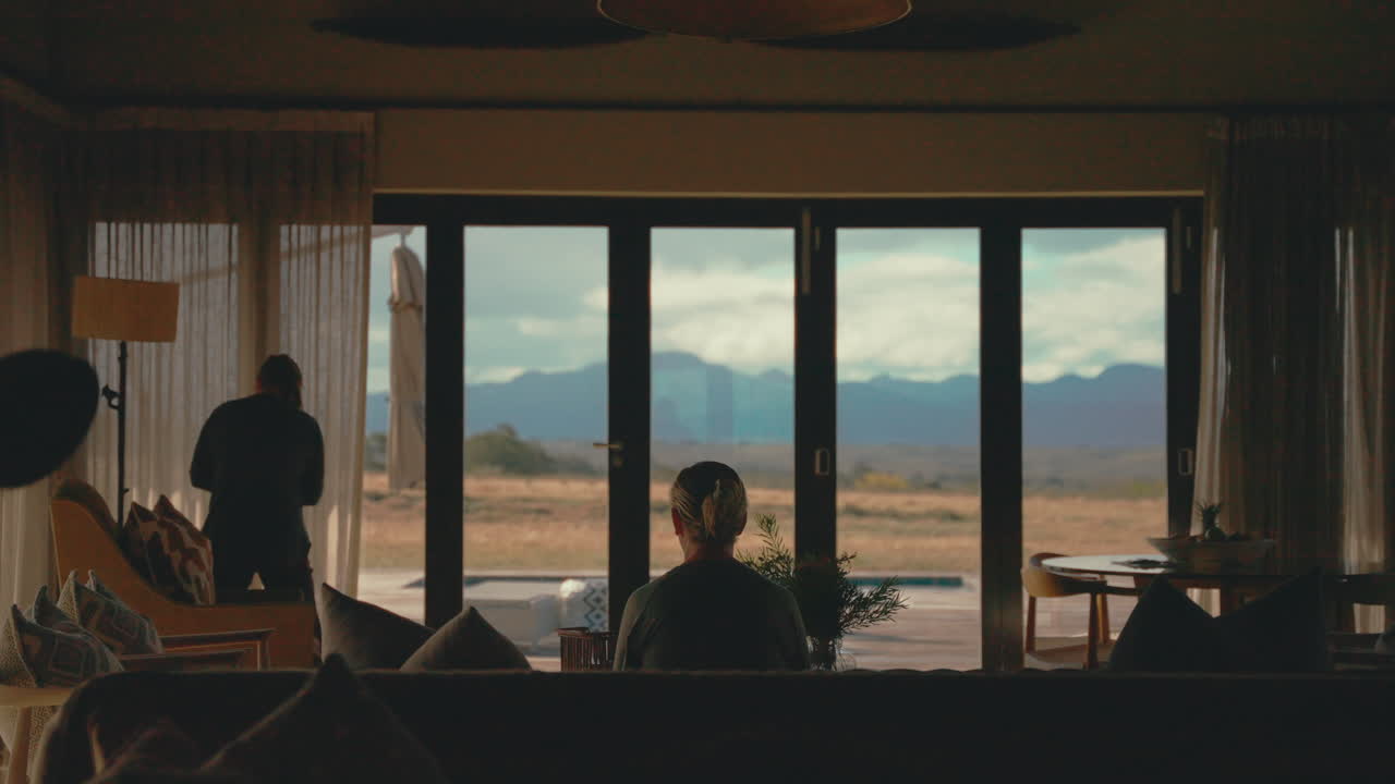 Woman sitting in scenic living room looking out large windows at mountainous landscape, static 4k