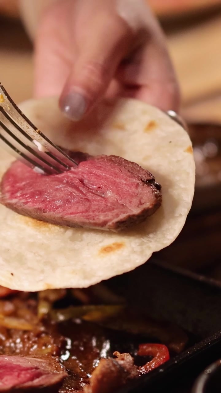 Tangan merangkai fajitas daging sapi dengan tortilla dan sayuran panggang di bawah pencahayaan hangat di sebuah restoran Bangkok.