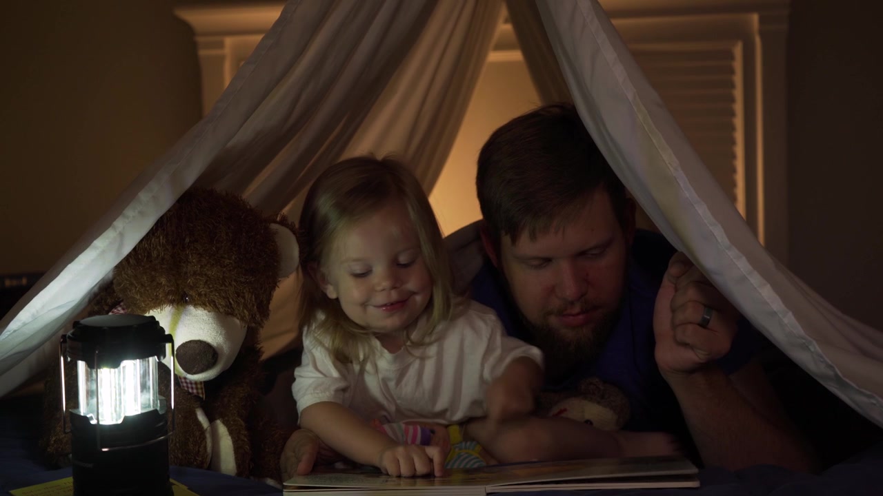 Father reading to toddler daughter at bedtime in blanket fort
