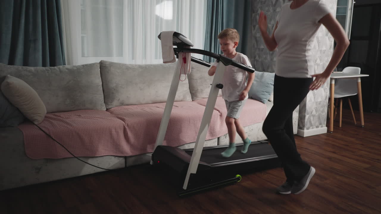 Young boy steps confidently on treadmill during home workout as mother in sportswear guides him with gentle gestures in cozy living room setup featuring sofa, curtains, and wooden floor