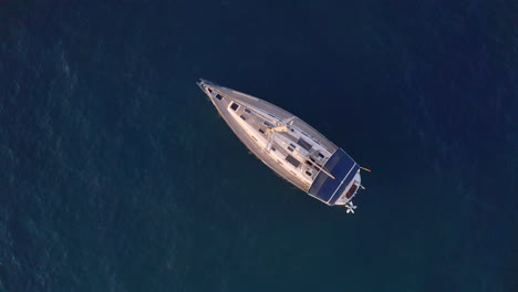 Overhead-4k-shot-of-a-luxurious-private-yacht-with-a-tall-mast-on-a-dark-blue-sea-anchoring-close-to-a-rocky-coast-on-a-sunny-summer-day