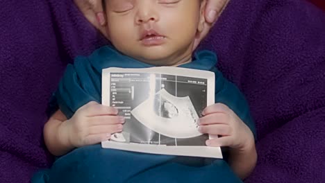 newborn baby holding ultrasound copy in hand and sleeping at mother palm in baby wrap