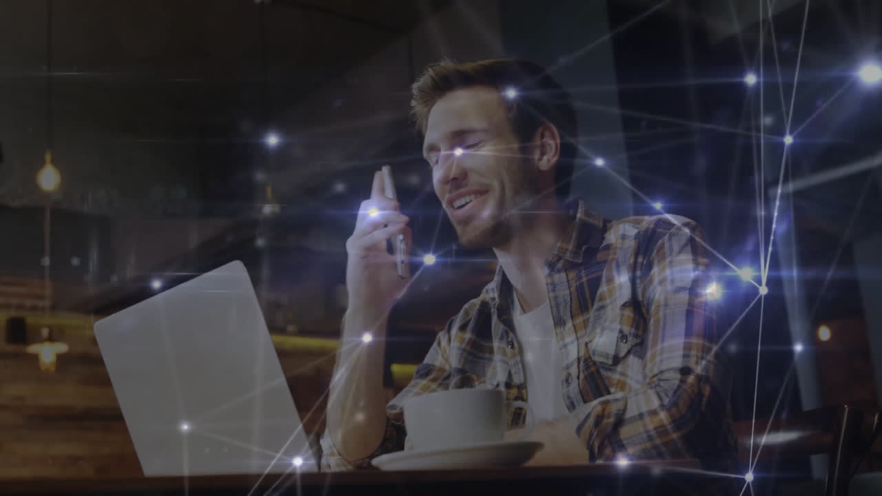 Remote worker typing on laptop holding smartphone in tech cafe, showing glowing network overlay