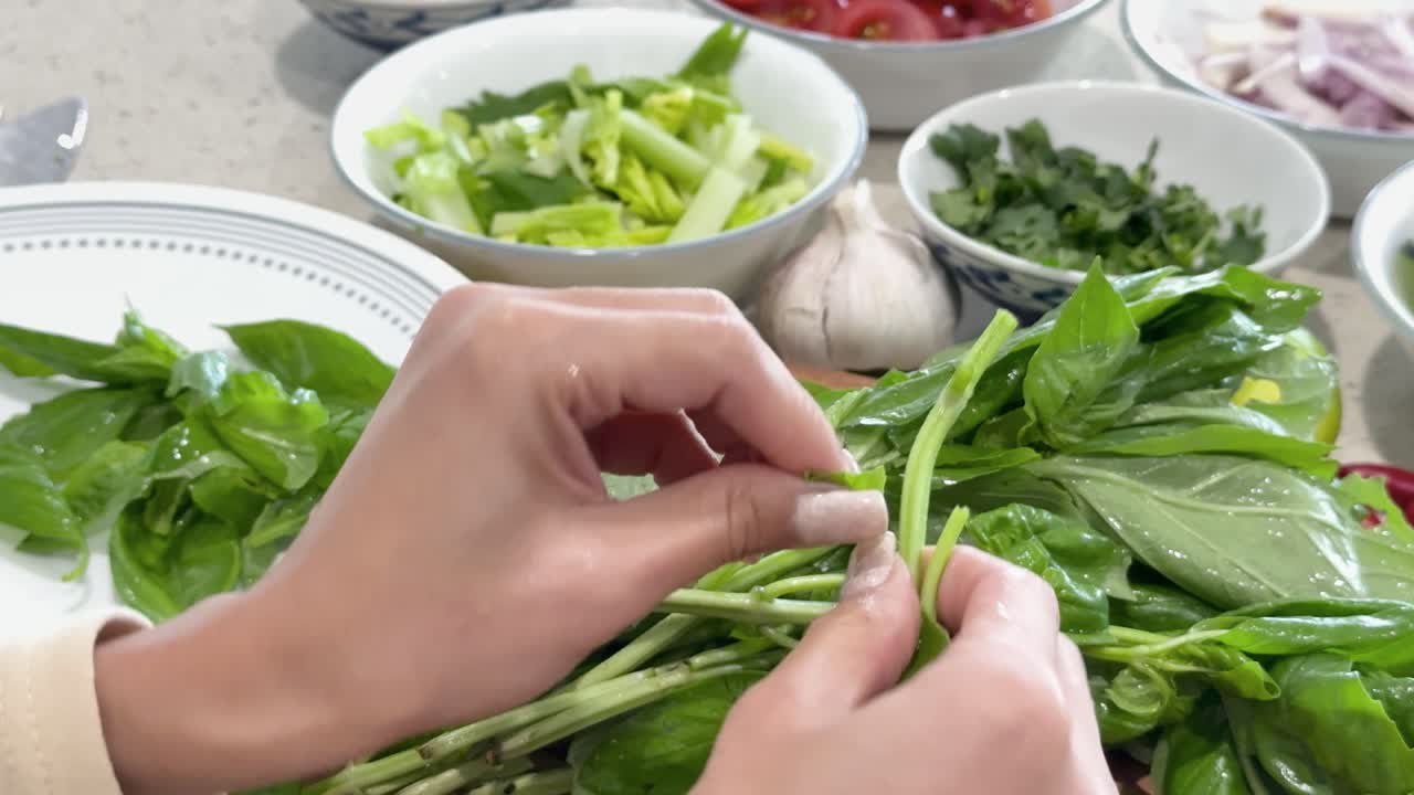 Close-up of hands picking basil leaves in a bright kitchen, surrounded by fresh vegetables and herbs, with soft natural lighting and steady camera