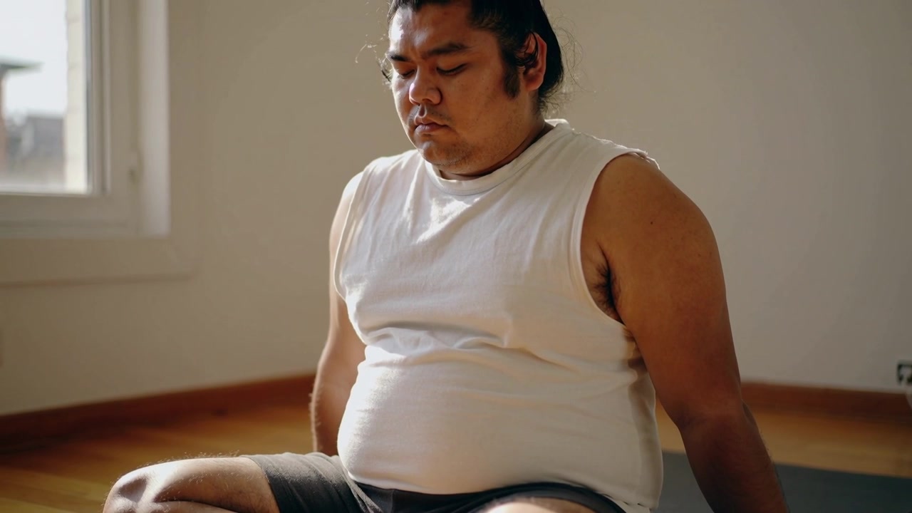Hombre asiático sentado con camisa y pantalones cortos, respirando y descansando en una esterilla junto a la ventana mientras entra la luz del sol, con un auricular.