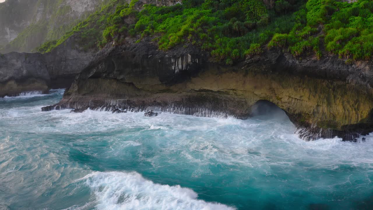 Stunning cinematic aerial view of waves of the ocean crashing on the exotic green and lush cliffs