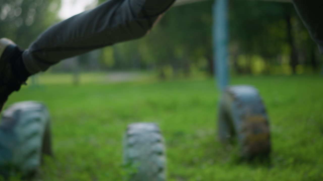 3개의 타이어를 가로질러 스프린트하는 누군가의 클로즈업 다리 뷰, 다른 타이어가 잔디에 배열되어 있으며, 배경에는 흐릿한 골포스트와 기둥이 있습니다.