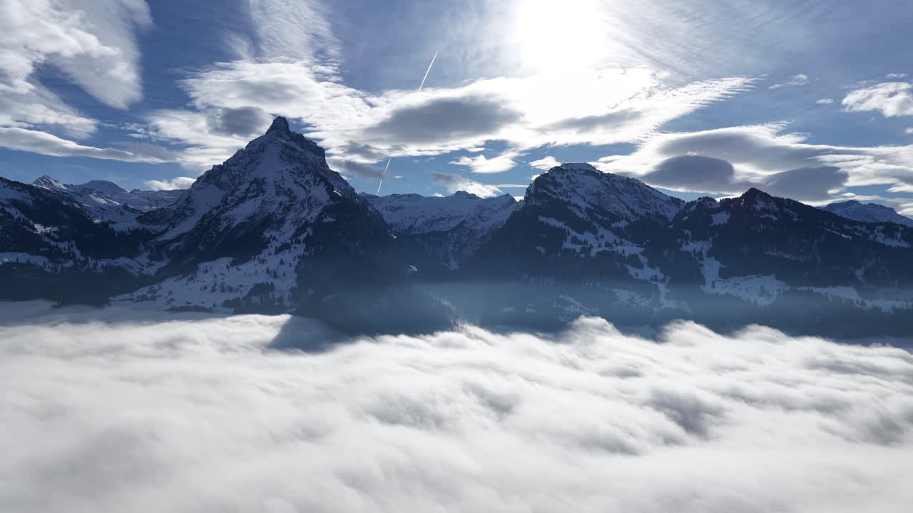 Drone view of snowy Swiss mountains above a dramatic cloud inversion during winter. Peaceful landscape featuring cliffs forest and soft morning light