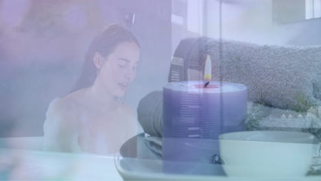 animation of scented candle and towels over caucasian woman in bath splashing face