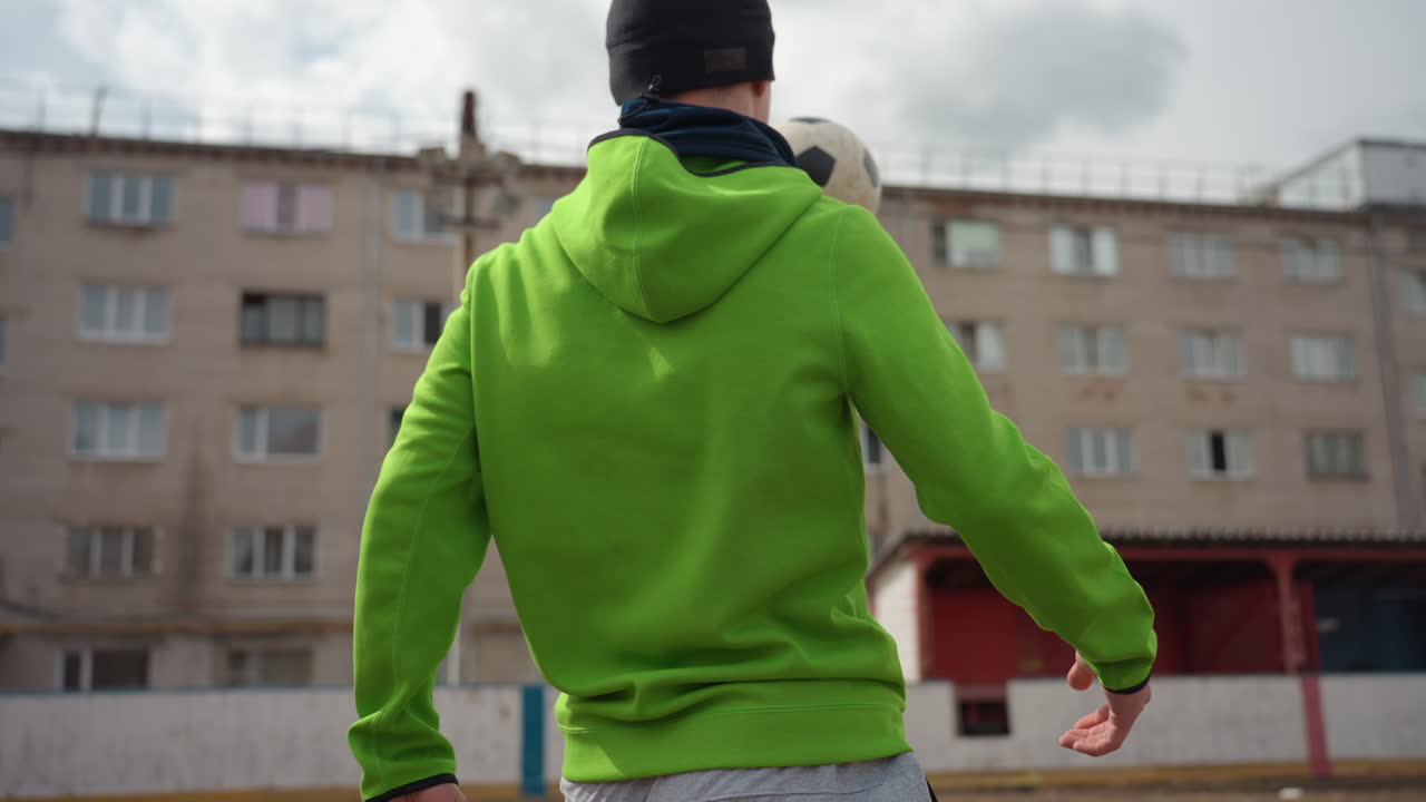 Крупный план: рука европейца контролирует футбольный мяч возле городского корта, рукав зеленой толстовки и детали ладони, сосредоточенное касание и точный расчет времени, фон с жилым районом и многоквартирными домами, атмосфера технических тренировок