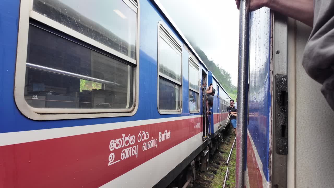 A passenger poses hanging out of a stationary blue and red train in overcast, rainy weather in Sri Lanka, capturing a daring photo opportunity.