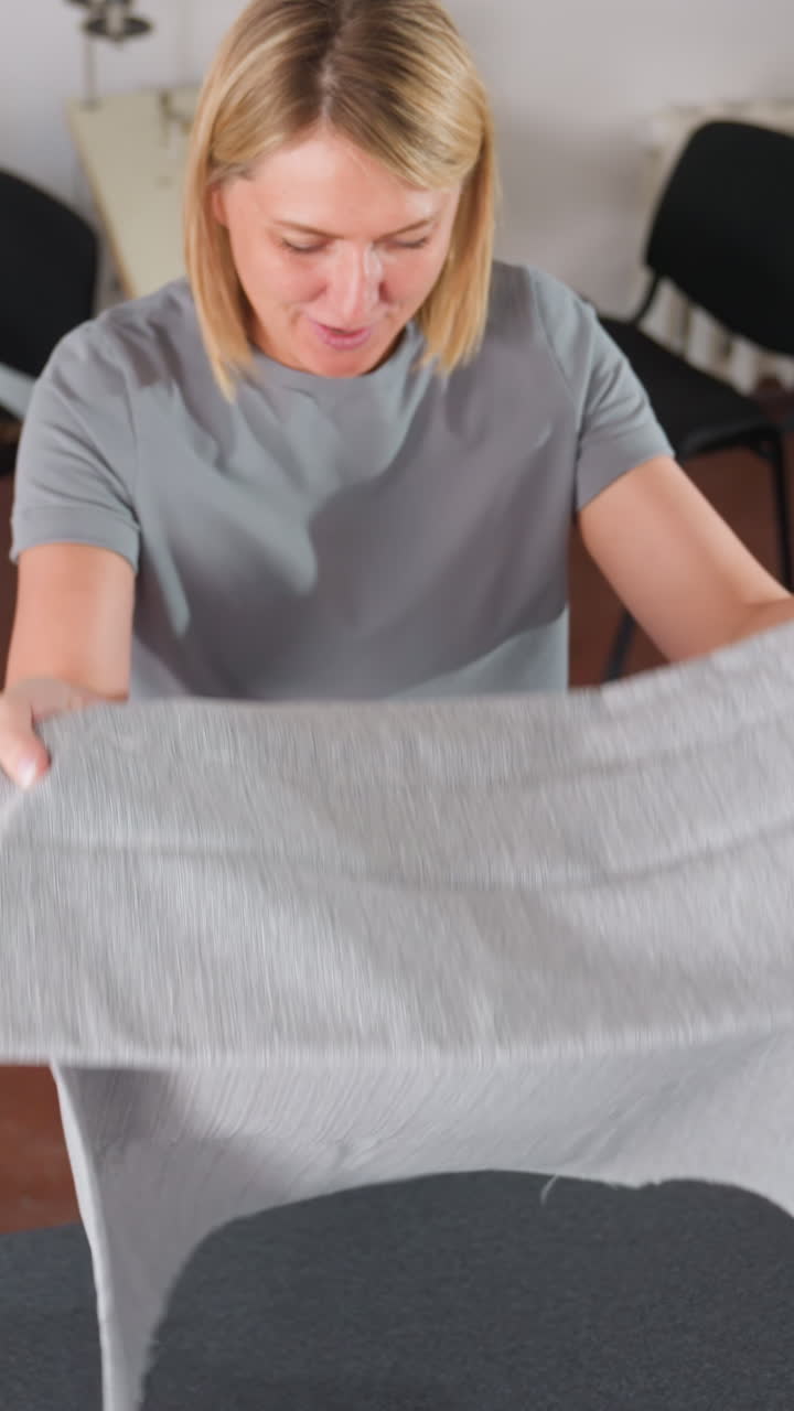 재단 테이블 위에 회색 원단을 펼쳐 패턴 용지를 정렬하고 원단을 매끄럽게 다듬으며 접힌 부분을 측정하고 도구를 사용하여 가장자리를 표시하는 여성 재봉사, 배경에는 산업용 재봉틀, 체계적인 모습