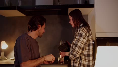 Happy-brunette-guy-with-stubble-in-a-gray-T-shirt-feeds-his-brunette-girlfriend-in-a-checkered-shirt-with-orange-slices-while-she-prepares-tea-in-the-kitchen-in-a-modern-apartment-in-the-evening