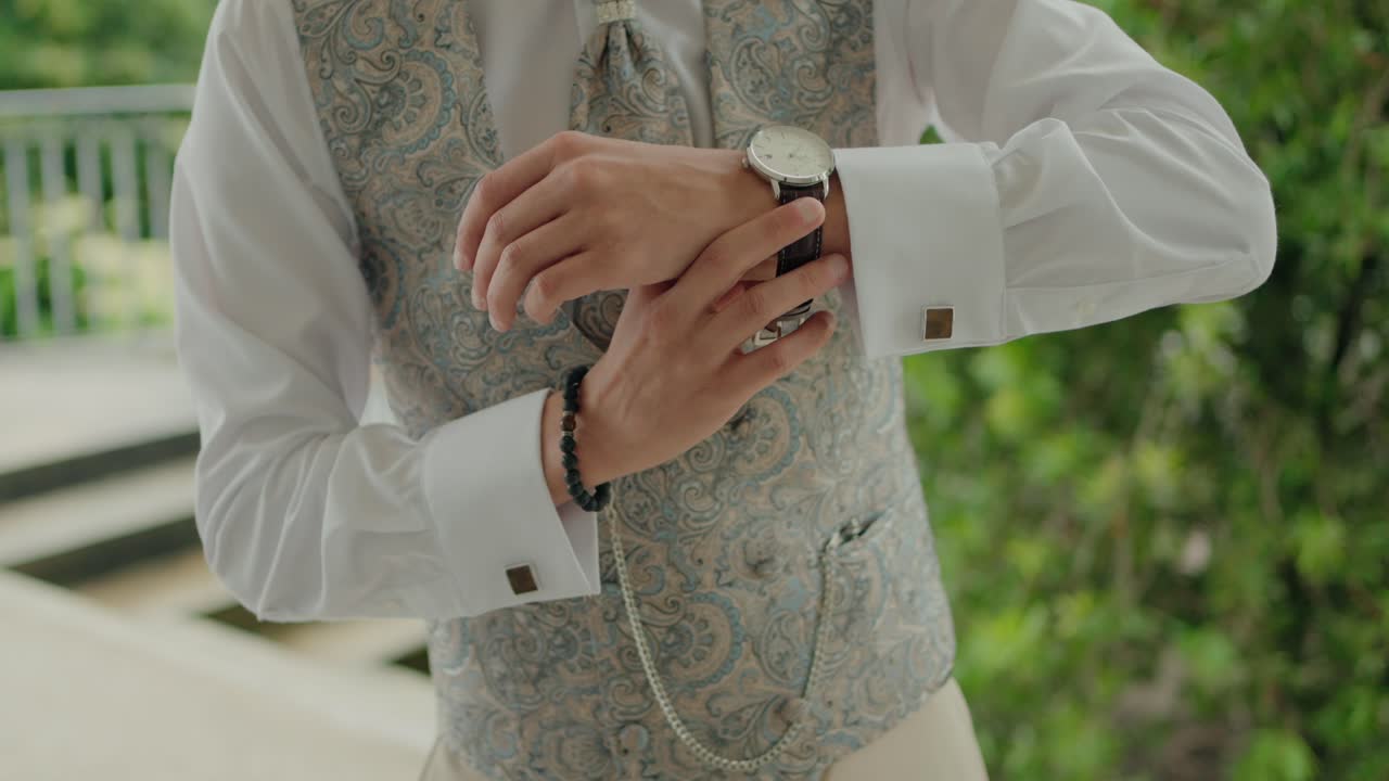 Groom adjusting his wristwatch in bright daylight, wearing a formal vest and shirt.