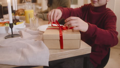 blond boy sitting at the table opens a gift, checks that it is a camera, and then hugs his mother