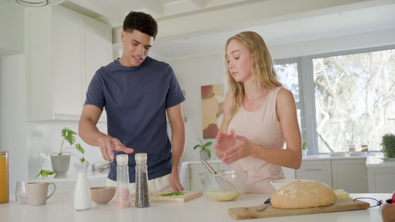 Casal cozinhando na ilha da cozinha, a mulher batendo ovos, o homem chegando para temperar a mistura com moedores.