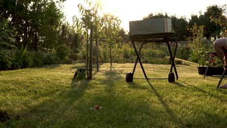 barbacoa con fuego en la naturaleza, parrilla en el jardín y un hombre sentado en una silla, un perro juega a la luz del sol y árboles en el jardín