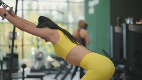 a young hispanic woman in a yellow tracksuit performs an exercise in a crossover pulls a steel rope from above to train her back and shoulders. a woman trains her back and shoulders in a gymnasium