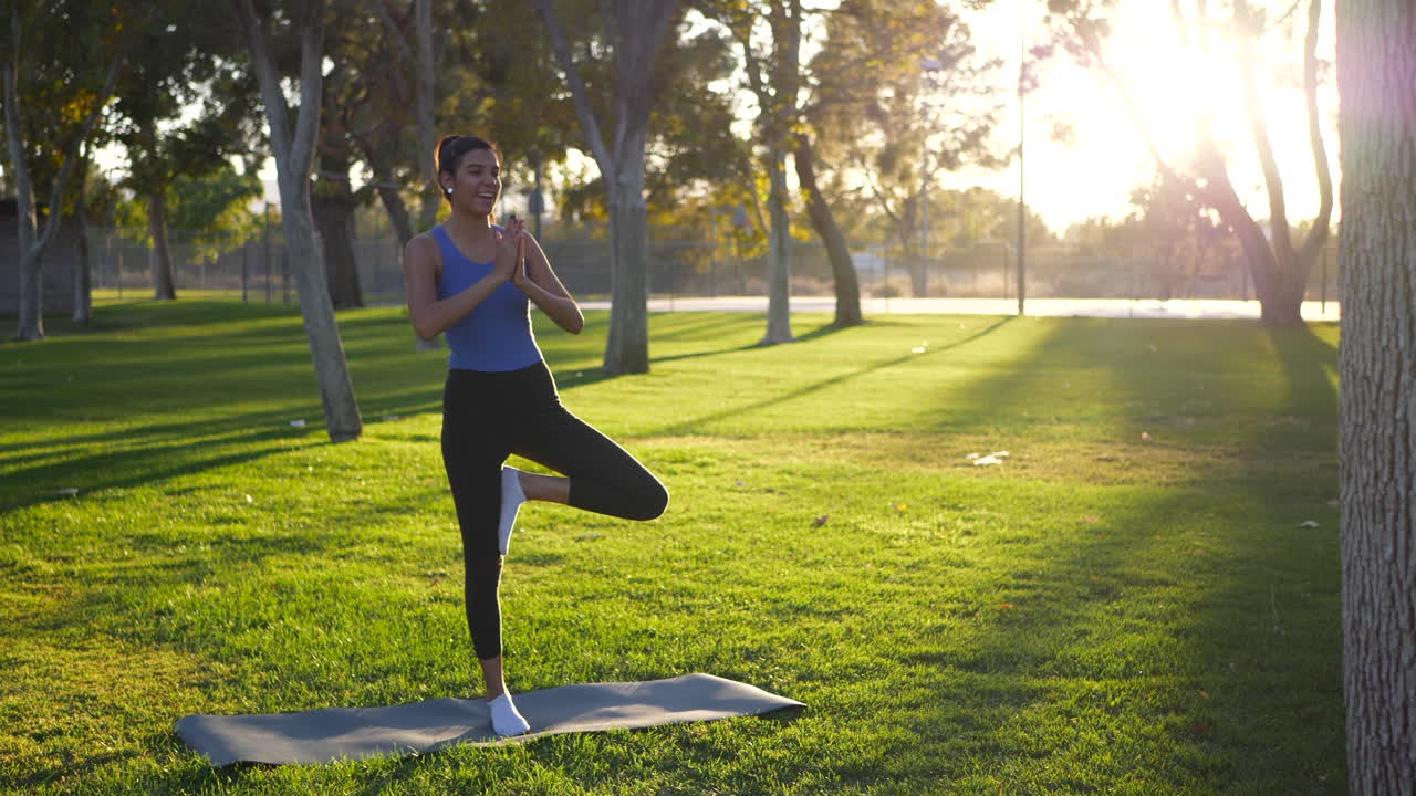 A beautiful young woman almost falling over and losing balance in a one legged prayer hands yoga pose on a mat in the park at sunrise