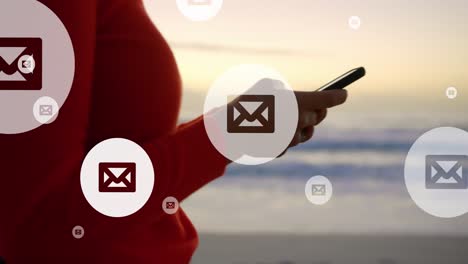 animación de iconos de correo sobre la sección media de una mujer biracial que usa un teléfono inteligente en la playa