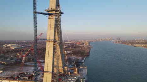 Construction-of-new-bridge-over-vast-river-with-Detroit-skyline-in-horizon,-aerial-view