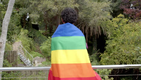 Standing on balcony, person wrapped in rainbow flag, enjoying nature view, copy space