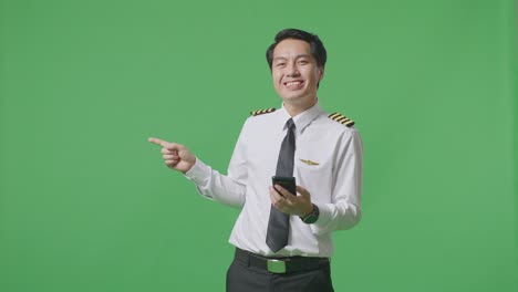 녹색 스크린 배경 스튜디오 에 서 있는 동안 스마트폰 을 사용 하고  을 가리키는 아시아 사람 조종사