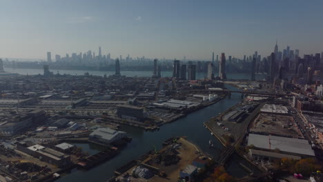 Fly-above-industrial-borough,-logistic-or-production-facilities-along-Newtown-Creek-water-transport-way.-Manhattan-business-skyscrapers-in-distance.-New-York-City,-USA