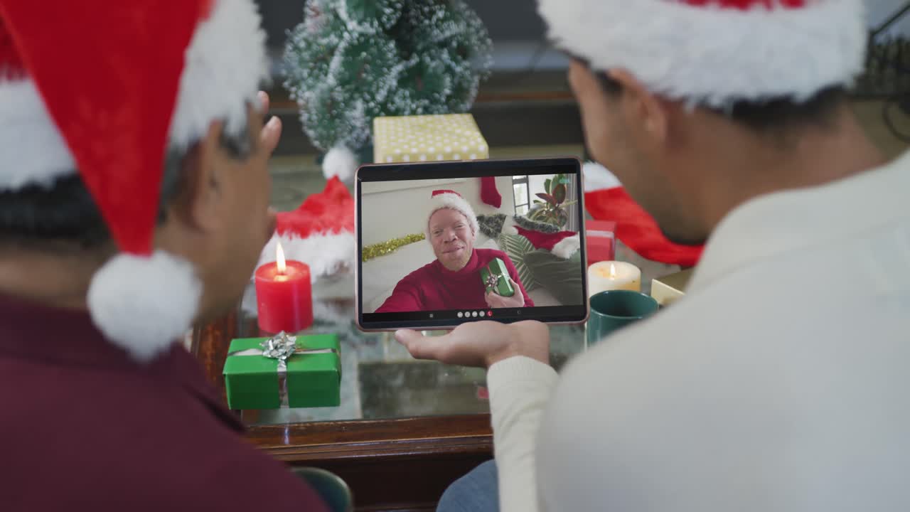 padre y hijo biraciales con sombreros de santa usando una tableta para una videollamada de navidad con un hombre en la pantalla